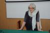 Starsza kobieta w siwych włosach i w okularach oparta rękami o stół stoi przy tablicy i głosi wykład.