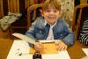Mała dziewczynka z gęsim piórem w prawej dłoni siedzi za stołem i własnoręcznie podpisuje kartkę papieru. Śmieje się do zdjęcia.