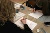 Dwie nastolatki w wieku licealnym siedzą przy biurku naprzeciw siebie i rysują na białych kartkach papieru za pomocą ołówka szkic kościoła zgodnie z oryginałem.