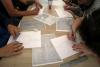 Grupa nastolatek w wieku licealnym siedzi przy stole naprzeciw siebie i rysuje na białych kartkach papieru za pomocą ołówka i linijek szkic kościoła i budynków zgodnie z oryginałem. Widać stół i dłonie uczestników warsztatów.