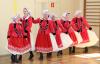 Sześć kobiet w czerwonych sukniach ludowych i białych czepkach z wyszywanymi wzorami na głowach występują na środku sali gimnastycznej prezentując taniec ludowy.