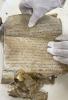 Dwie dłonie w białych rękawiczkach trzymające rozwinięty rulon papieru z zapisanym ręcznie tekstem w języku niemieckim. Dolna część dokumentu poważnie uszkodzona.